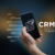 A person's hand holding a smartphone with the letters CRM displayed on the screen. The image highlights the benefits of using a CRM system, such as improved customer relationship management, increased sales and efficiency, and enhanced data analysis capabilities.